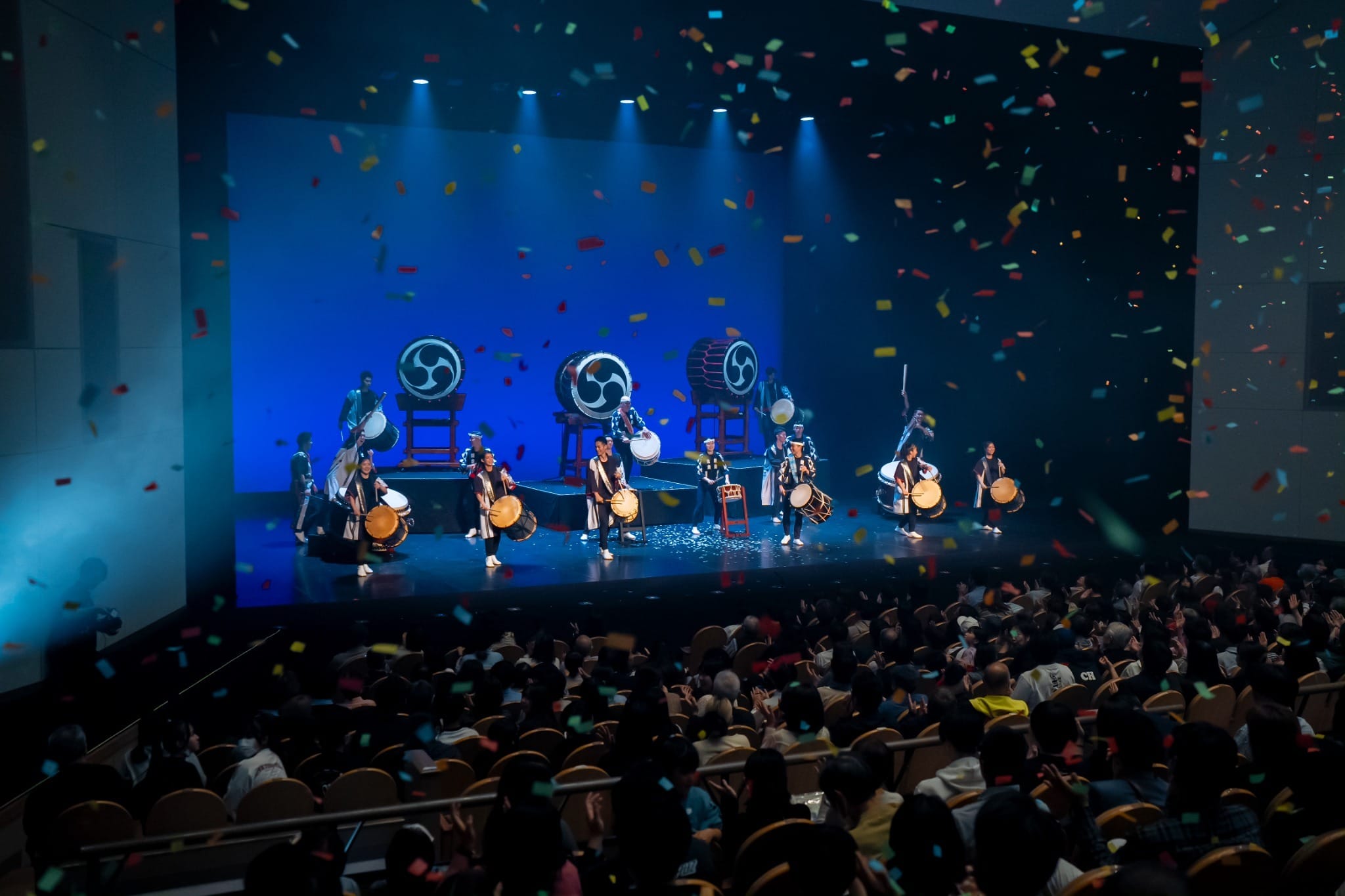大ホールの舞台で和太鼓を演奏する出演者と、客席を埋める観客。舞台上に紙吹雪が舞う公演の一場面